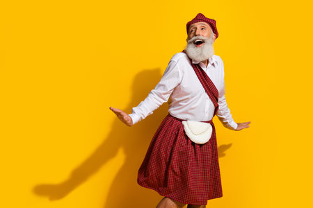 Elderly Man in Traditional Scottish Attire Joyfully Posing on a Yellow Background with Expressive Gesturesの写真素材