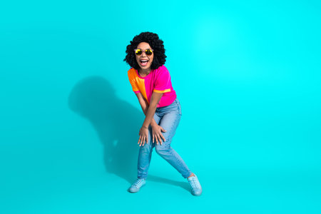 Joyful woman in casual clothing and sunglass standing against a vibrant turquoise background