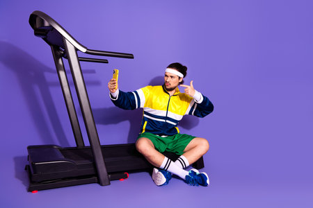 Young athlete in retro activewear taking a selfie near treadmill against vibrant purple background,...