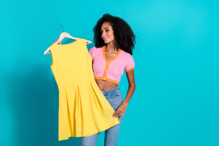 Young woman holding a bright yellow dress against a vibrant turquoise background, smiling confidently in fashionable casual wear.の写真素材