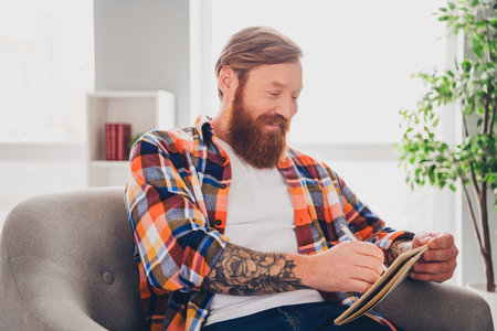 Middle aged bearded man enjoying leisure time indoors, wearing casual checkered shirt in bright modern living room settingの写真素材