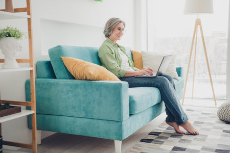 Senior woman sitting on an elegant sofa, using a laptop in a bright and cozy home interior, exuding happinessの写真素材