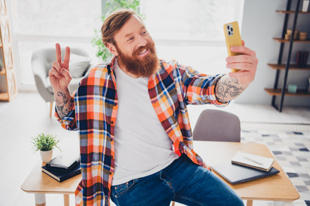 Smiling bearded man in plaid shirt taking a selfie while posing in a bright and cozy living room environmentの写真素材