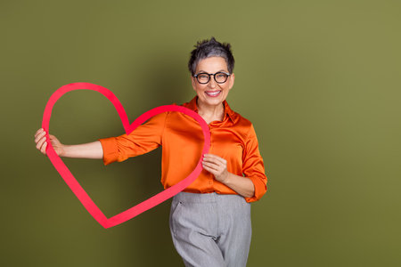 Mature woman posing with a red heart symbol while smiling in elegant attire, standing against a khaki backgroundの写真素材