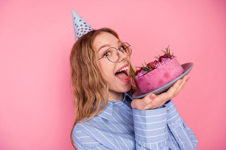 Smiling young woman holding a colorful cake, wearing glasses and party hat, against a pink background, celebrating with joyの写真素材