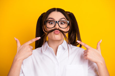 Young brunette student with glasses and playful expression in casual white shirt against vibrant yellow backgroundの写真素材
