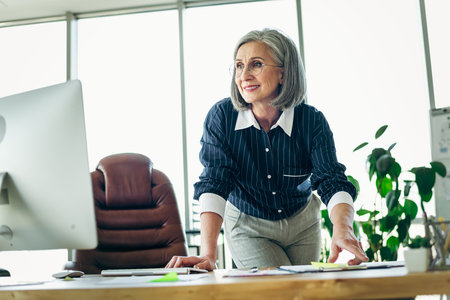 Confident mature businesswoman standing in a modern office leaning on a desk with a professional and engaging demeanorの写真素材