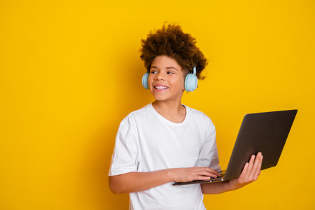 Smiling boy with headphones holding a laptop against a vibrant yellow background, embodying modern...