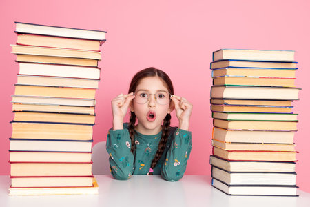Excited schoolgirl with glasses between stacks of book, expressing amazement against a vibrant pink...