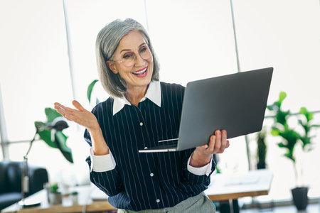 Smiling mature businesswoman using a laptop in a modern office setting, looking professional and confident in formal attireの写真素材