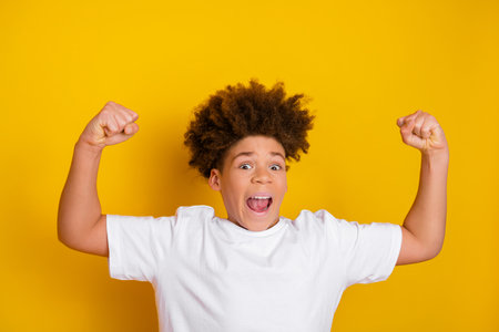 Excited young boy with curly hair smiles and poses joyfully in a white t-shirt against a vibrant yellow backdropの写真素材