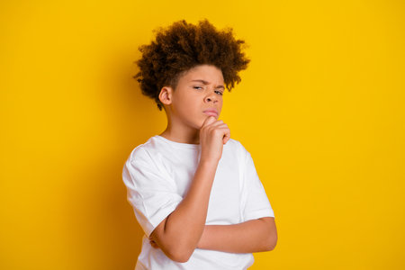 Confident young boy considers decision while displaying thoughtful expression against vibrant yellow backgroundの写真素材