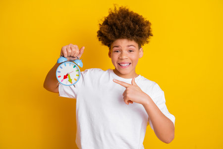 Smiling young boy holding an alarm clock and pointing at it on a vibrant yellow background, promoting time awarenessの写真素材