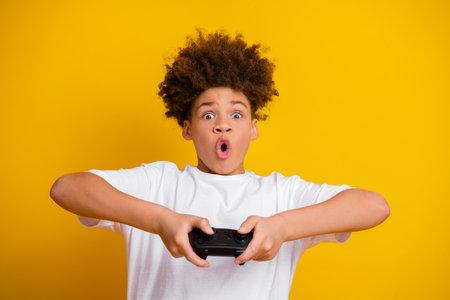 Excited young boy with curly hair holds game controller, expressing surprise against bright yellow backgroundの写真素材