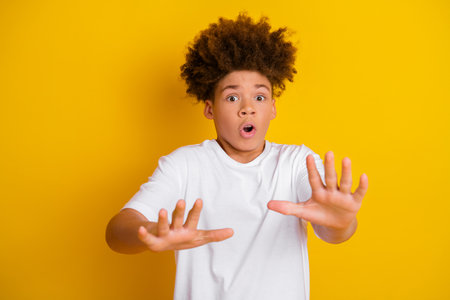 Surprised young boy expressing astonishment with hands raised against vibrant yellow backgroundの写真素材