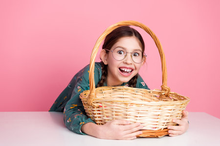 Adorable young girl with braids and glasses smiling brightly, holding a woven basket against a vibrant pink background.の写真素材