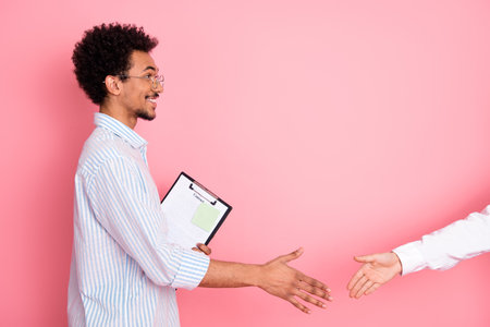 Young male professional in a casual setting offering a handshake, a clipboard in hand, positioned against a pink backgroundの写真素材