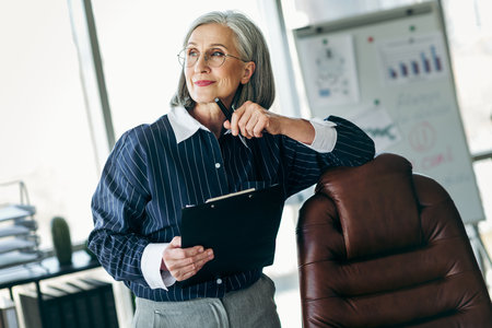 Confident professional mature businesswoman in stylish formal blouse holding clipboard while thinking in office settingの写真素材