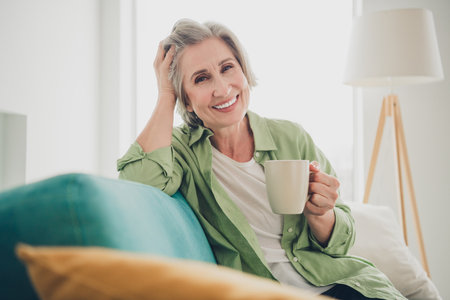 Smiling senior woman relaxing at home with a cup of tea, sitting on a sofa in a cozy living roomの写真素材