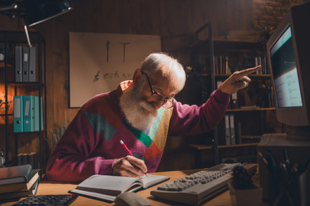 Elderly Man Working at Desk in Cozy Office Setting Using a Vintage Computer and Taking Notes, Embracing Technologyの写真素材