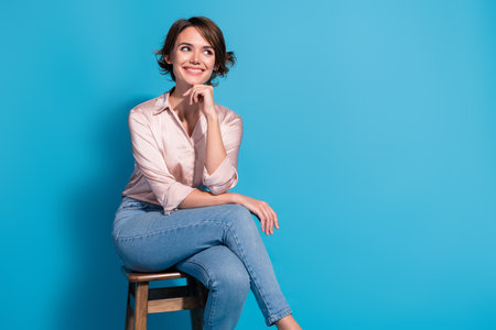 Young woman in satin pink blouse sitting on stool against blue background looking thoughtful and smiling, conveying elegance and styleの写真素材