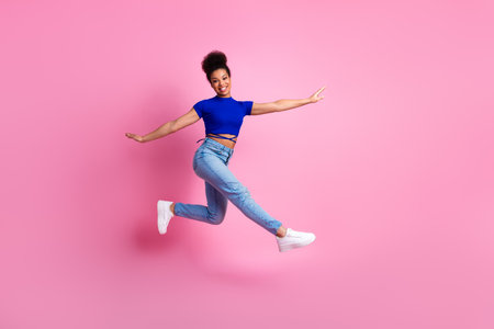 Excited young woman in casual stylish attire jumping enthusiastically against a vibrant pink...