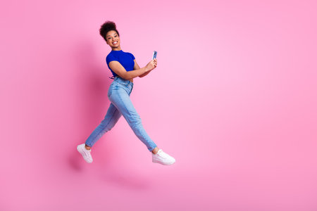Joyful young woman in casual blue and denim outfit jumping with phone on pink background, expressing...