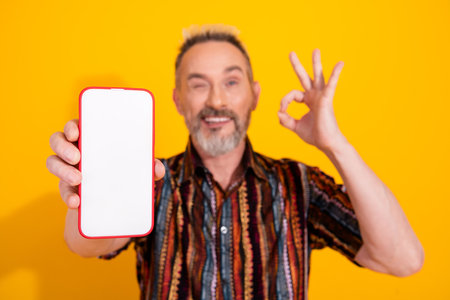 Smiling mature man with a smartphone shows okay gesture against a bright yellow background dressed...