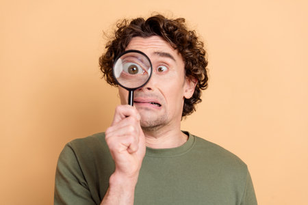 Young man with curly hair holding magnifying glass displaying a surprised expression against beige...