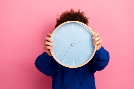 Young boy holding a modern clock in front of his face against a vibrant pink background, expressing time management and playful vibesの写真素材