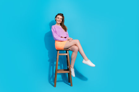 Young woman sitting on a wooden stool in bright casual summer wear against a blue studio background, smiling confidentlyの写真素材