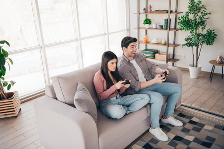 Young couple enjoying indoor leisure time playing video games together in a cozy living room with a modern decor and natural daylightの写真素材