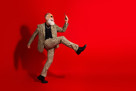 Elderly man in leopard print suit, holding smartphone, dancing joyfully on bright red background, combining elegance, style, and livelinessの写真素材