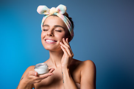 Radiant young woman applying skincare cream with natural beauty on blue backgroundの写真素材