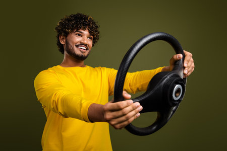 Cheerful young man holding a steering wheel in front of a khaki background wearing a trendy yellow...