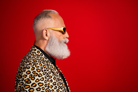 Stylish senior man in leopard print jacket and trendy glasses posing against a vibrant red backdrop showcasing confidence and charismaの写真素材