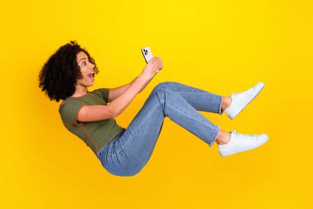 Cheerful young woman with curly hair looking at her smartphone while floating against a vibrant...