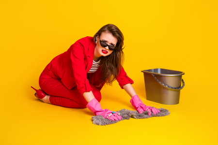 Stylish young woman scrubbing floor while wearing fashionable red suit and gloves against brightly...
