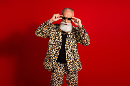 Stylish senior man in leopard print suit and sunglass posing confidently against vibrant red backgroundの写真素材