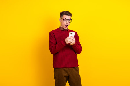 Handsome Young Handsome Man in Stylish Red Sweater Looking Surprised at Smartphone Over Yellow...
