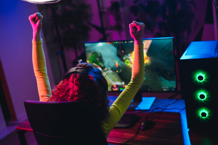Charming girl celebrating victory while playing an online video game at home with neon lights during the nightの写真素材