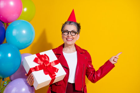 Senior woman in a red leather jacket celebrating with gift box and colorful balloons on yellow backgroundの写真素材