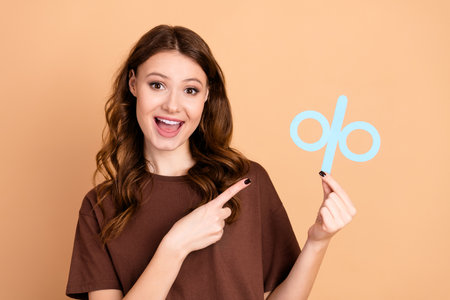 Joyful Young Woman Pointing to a Blue Percent Sign Symbol on a Beige Background Demonstrating...