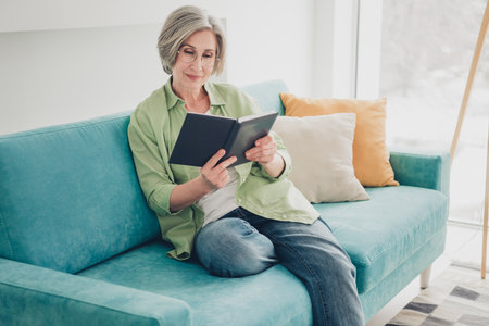 Mature Woman Relaxing on a Comfortable Sofa Reading a Book at Home in a Well-Lit Living Roomの写真素材