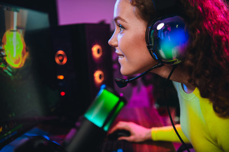 Charming girl gamer engaged in streaming with headset and microphone illuminated by neon lights for...