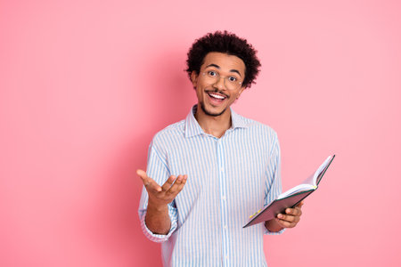 Confident young man holding an open book and gesturing against a pink background symbolizing learning and communication.の写真素材