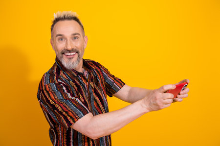 Charismatic mature man with blond hair wearing a trendy print shirt smiling and holding a red phone on yellow backgroundの写真素材