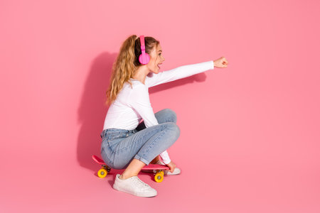 Young woman on a skateboard wearing headphones, enjoying leisure and showcasing vibrant energy against a pink backdropの写真素材