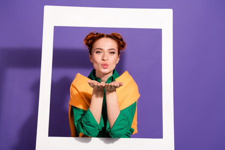 Lovely woman with red hair and colorful fashion blowing a kiss framed by a white border against a purple backdropの写真素材