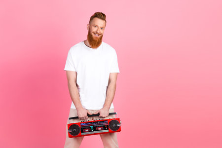 Man with red beard holding a retro boombox in a casual white shirt against a vibrant pink backdrop, stylishly posedの写真素材